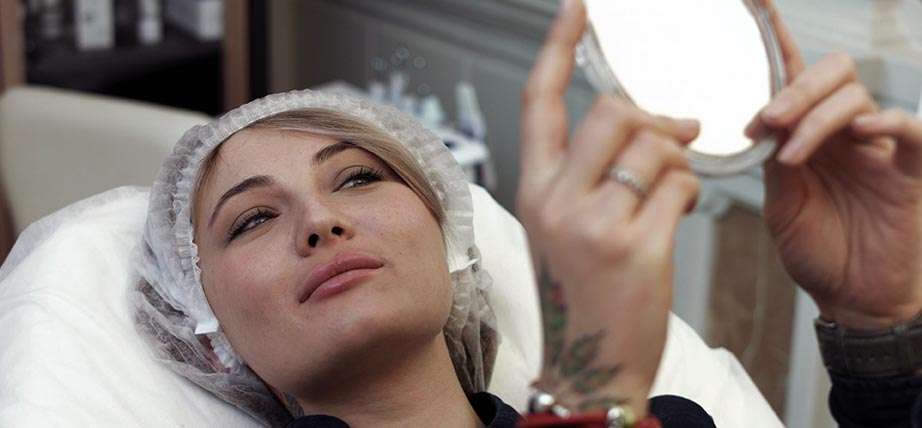 A woman happily examines her reflection in the mirror after getting fillers.