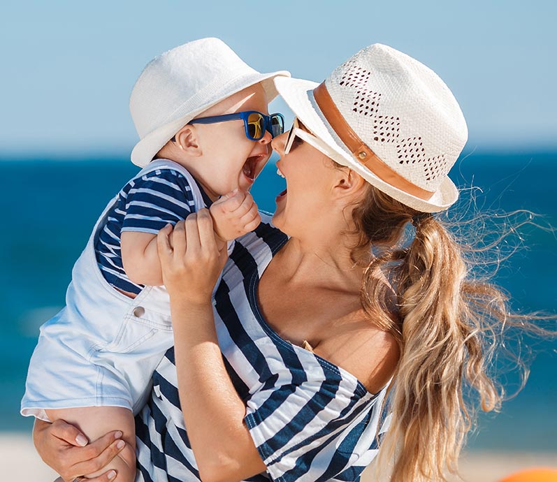 A happy mommy makeover patient holds her infant son at the beach.